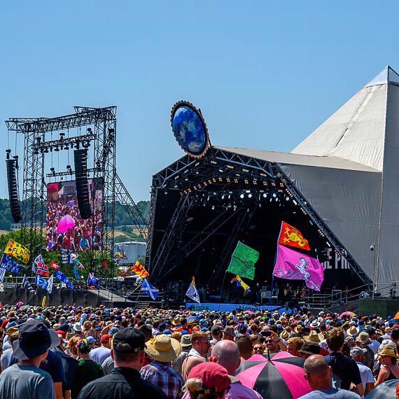 "A Glastonbury festival stage with thousands of people in attendance on a sunny day"