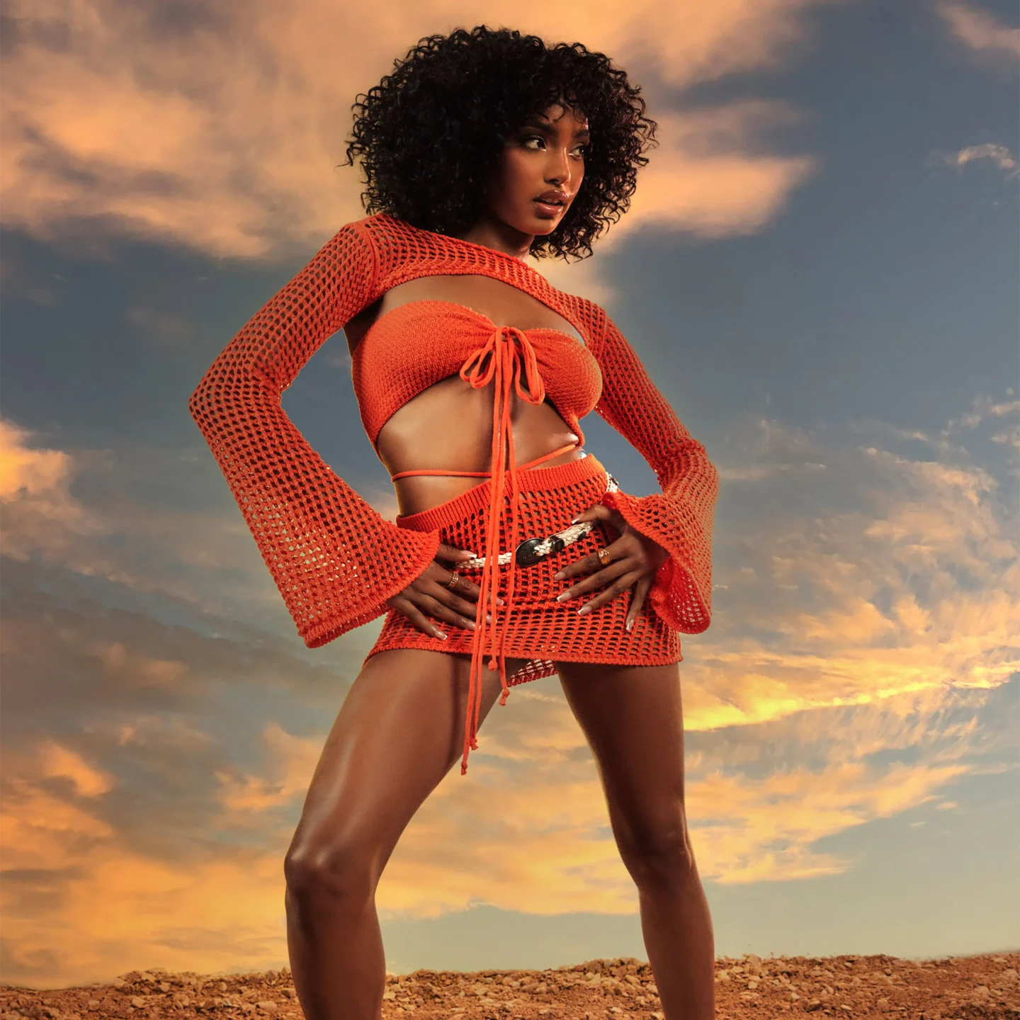 An orange crochet co-ord set including a crochet bandeau bralet that ties at the front, a crochet mini skirt with strappy waist details and a long bolero sleeve .